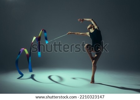 Gymnast child girl lit by light in black bodysuit performs gymnastic exercises with ribbon in her hand on dark background.