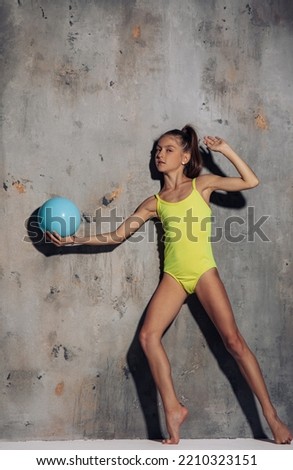Gymnast child girl in yellow bodysuit performs gymnastic exercises with ball in her hand on gray background.