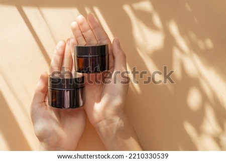 Two jars of moisturizer in women's hands. Amber glass bottles with gel, shampoo, cosmetic, close-up on beige background in sunlight. Top view, flat lay, copy space Royalty-Free Stock Photo #2210330539