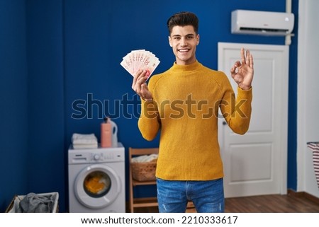 Young hispanic man at laundry room holding shekels doing ok sign with fingers, smiling friendly gesturing excellent symbol 