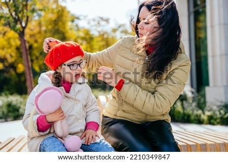 Brunette caucasian mother with loving corrects beret her little daughter. They are sits on bench outdoor. Enjoying Spending Time Together.
