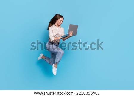 Full body photo of optimistic millennial lady jump look laptop wear shirt jeans boots isolated on blue color background