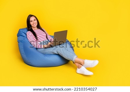Full size photo of positive cheerful girl striped shirt jeans sit in pouf with laptop chatting texting isolated on yellow color background