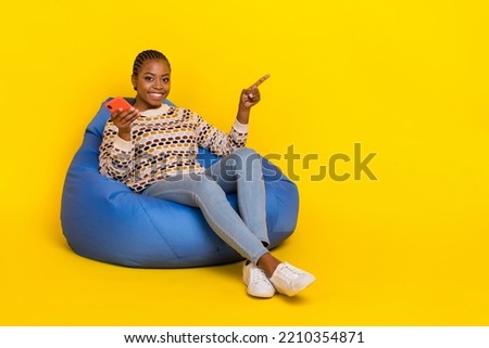 Full size photo of attractive positive lady sit comfy beanbag hold telephone direct finger empty space isolated on yellow color background