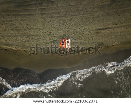 Aerial view from a drone of two young slender women in a Santa Claus hat lying on a sandy beach of the Black Sea with rolling waves at sunset. Winter holidays at sea