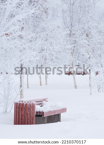 A snow-covered park bench. The trees are covered with fluffy white snow. Vertical photo. Snow day.