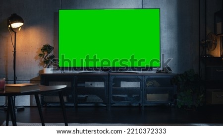 Stylish Loft Apartment Interior with TV Set with Green Screen Mock Up Display Standing on Television Stand. Empty Cozy Living Room of Spacious Flat with Chromakey Placeholder on Monitor. Royalty-Free Stock Photo #2210372333