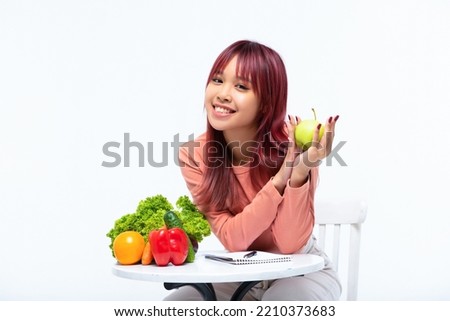 Beautiful Asian young woman nutritionist smiling sitiing and holding green apple. Table with vegetables.  Natural and meal concept. Healthy food and diet. Isolated on white background. Copy space