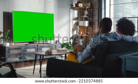 Black Couple Sitting At Home in the Living Room During the Day and Watching Green Screen Chroma Key on Television Set, Relaxing on a Couch. Weekend Activities and Mainstream Media Concept.