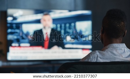 Man Watches Late Night News Show on TV While Sitting on a Couch at Home in the Evening. Presenter Talks on TV. Cozy and Stylish Loft Living Room with Warm Lights. Over the Shoulder Shot. Royalty-Free Stock Photo #2210377307