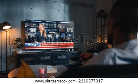 Over the Shoulder View of Man Watching TV News, Sitting on Couch in Stylish Loft Living Room. TV Presenter Telling Breaking News About Construction Industry. Home with Cozy Interior Concept. Royalty-Free Stock Photo #2210377317