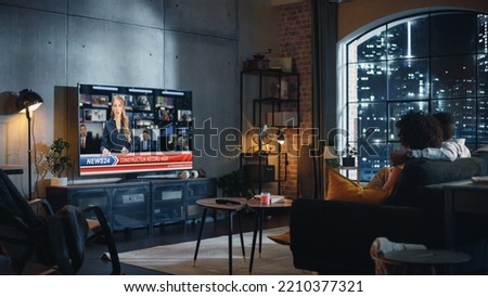 Couple Watches Evening News on TV While Sitting on a Couch at Home in the Night. Two Smart People Politely and in Civil Manner Discuss Important Political Issues of the Day. Back View. Royalty-Free Stock Photo #2210377321