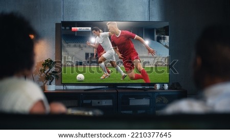 Black Couple Watches Professional Soccer Match on TV, Sitting on a Couch at Home in the Evening. Boyfriend and Girlfriend Football Fans Watch Sports. Back View Out of Focus Close Up Shot at Night. Royalty-Free Stock Photo #2210377645