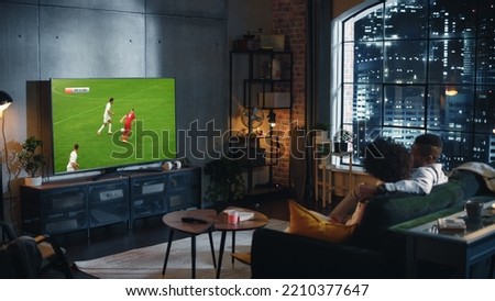 Couple of African American Soccer Fans Relax on a Couch, Watch a Sports Match at Night at Home in Stylish Loft Apartment. Young Man and Woman Enjoying the Game of Favorite Football Club. Royalty-Free Stock Photo #2210377647