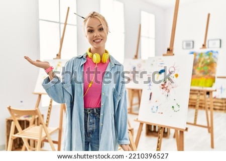 Young caucasian girl at art studio smiling cheerful presenting and pointing with palm of hand looking at the camera. 
