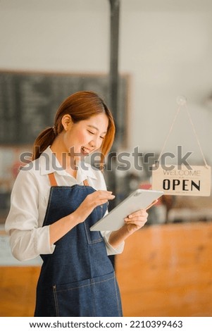 A younger shopkeeper standing in front of her shop. Open a shop. A beautiful Asian woman in an apron holds a tablet and stands in front of a cafe door with an open sign. business owner startup ideas