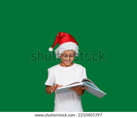 Boy in Santa Claus hat laughs and looks into an open book, which he holds with his hands. Green background with space for text. Selective focus. Picture for articles and advertisements about children.
