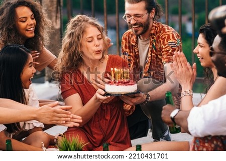 The multicultural friends celebrate a birthday. A man holds a cake with candles while a woman blows the candles. The rest of the group is singing the song and applauding.