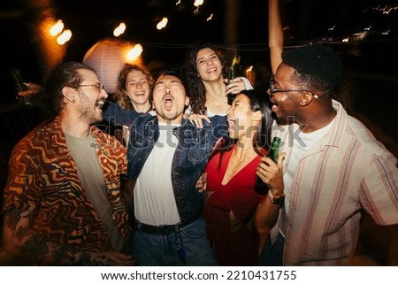 A group of drunk multiracial friends is having fun at the night open-air party. They are singing, drinking beer, and having a good time together. Royalty-Free Stock Photo #2210431755