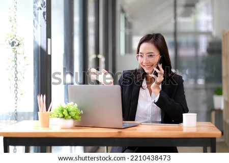 Happy businesswoman sitting at desk behind her laptop and talking with somebody on her mobile phone while working at office. Royalty-Free Stock Photo #2210438473