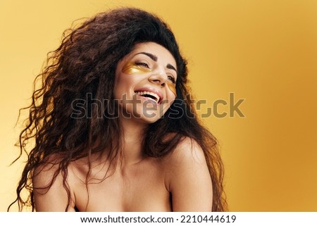Happy pretty curly Latin lady in gold hydrogel patches under eye enjoy flawless skin smiling aside posing isolated over yellow background. Cosmetic product ad Natural beauty concept Studio portrait