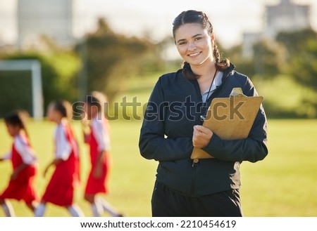 Soccer field, woman coach with and girl team training on grass in background. Sports, youth development and teamwork, a happy young female volunteer coaching football team with clipboard from Brazil. Royalty-Free Stock Photo #2210454619