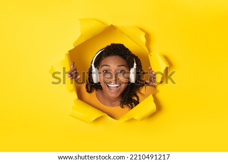 Joyful Black Lady Wearing Wireless Headphones Listening To Music Smiling Looking Through Hole In Torn Yellow Paper, Advertising Musical Application. Female Enjoying Favorite Song