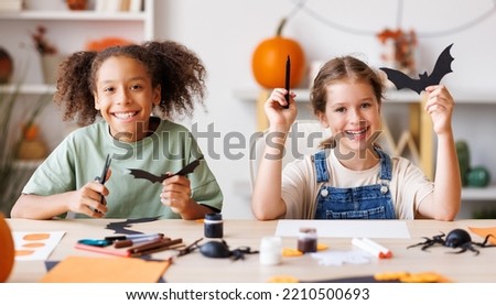 Happy multinational children girls making Halloween home decorations together, kids painting pumpkins and making paper cuttings