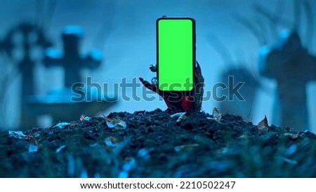 Zombie hand rising up smartphone with green screen out of grave. Holiday event halloween concept. Royalty-Free Stock Photo #2210502247