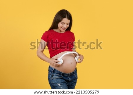 Happy Young Pregnant Woman Holding Wireless Headphones Near Belly, Smiling Expectant Female In Casual Clothes Playing Music For Baby In Womb While Standing Isolated On Yellow Background, Copy Space Royalty-Free Stock Photo #2210505279