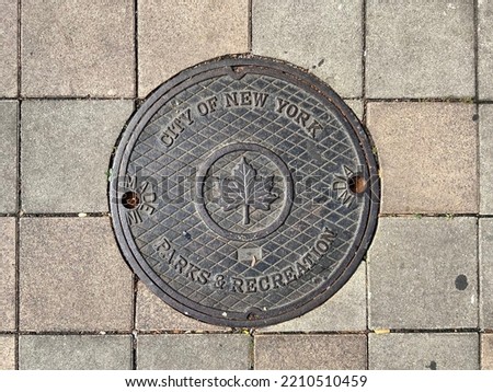 Close-up of a round metal manhole cover that reads "City of New York Parks and Recreation" with the leaf logo on the sidewalk in New York City