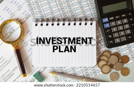 On the table are reports, diagrams, a pen, a magnifying glass and a white notepad with INVESTMENT PLAN text. Business concept.