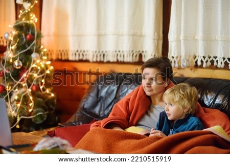 Young grandmother and cute grandchild are sitting on sofa in mountain chalet and watching Christmas movie together. Family leisure during Xmas. Winter holidays on cozy mountain chalet in European Alps