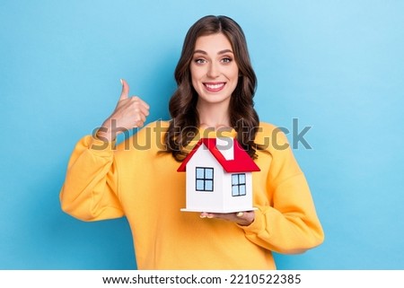 Photo portrait of charming young woman show thumb up hold small house dressed stylish yellow garment isolated on blue color background