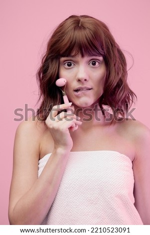 a happy woman, standing on a pink background with pink lighting from the side, with beautiful, styled hair, doing a facial massage with a pink roller, thoughtfully biting her lower lip. Vertical photo