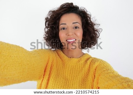 Photo of young beautiful brunette woman with curly short hair wearing yellow sweater over white wall do selfie
