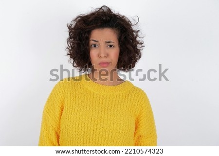 Displeased young beautiful brunette woman with curly short hair wearing yellow sweater over white wall frowns face feels unhappy has some problems. Negative emotions and feelings concept