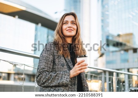 Young smiling woman in coat with paper coffee cup in evening city street Royalty-Free Stock Photo #2210588391