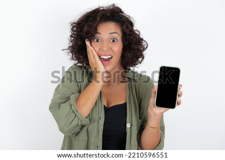 young beautiful woman with curly short hair wearing green overshirt over white wall hold hand modern technology use touch face palm astonished impressed scream wow omg unbelievable unexpected