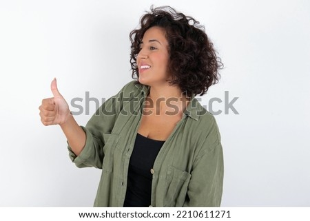 young beautiful woman with curly short hair wearing green overshirt over white wall Looking proud, smiling doing thumbs up gesture to the side. Good job!