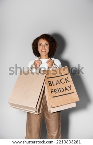 positive young african american woman holding shopping bags with black friday lettering on grey