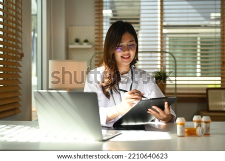 Portrait of female doctor wearing white coat using laptop and filling medical form at her workplace