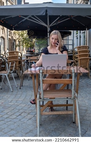 Attractive young blonde business woman using her laptop while calling with her smartphone, sitting outdoors in a European city at the typical Belgian cafe terrace. High quality photo