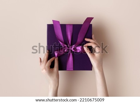 Female hands with festive minimalistic manicure tie a bow on violet gift box. Surprise preparation. Christmas Eve. Xmas and New Year postcard. Black Friday sales, Birthday celebration party concept