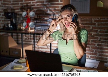 Young beautiful woman working at the office at night speaking on the phone doing peace symbol with fingers over face, smiling cheerful showing victory 