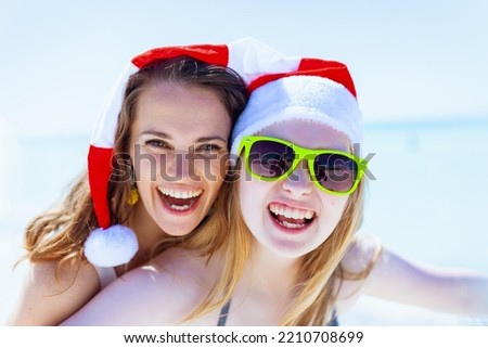 Portrait of smiling modern mother and teenage daughter in santa hat at the beach in beachwear having fun time.