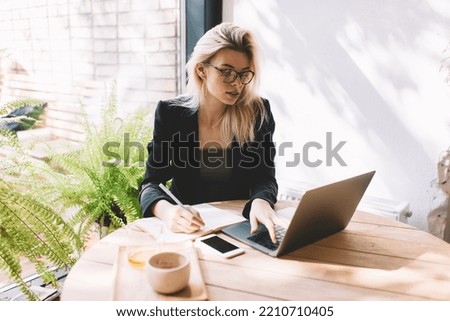 Caucasian female journalist with notepad using laptop application for planning content publications and browsing social networks, skilled woman in eyeglasses connection to 4g wireless on netbook