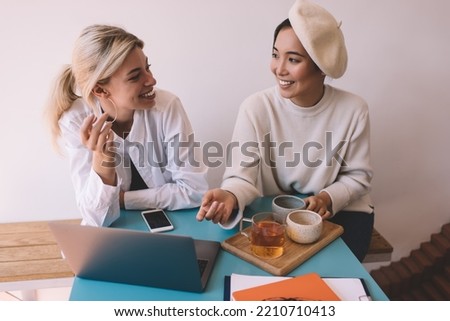 Joyful female bloggers with tea and caffeine beverages smiling during social communication in cafe interior, happy Asian and Caucaisan women talking about web design on netbook technology
