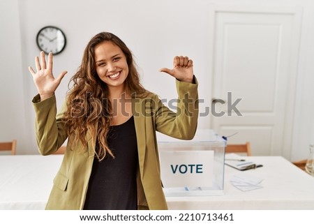 Beautiful hispanic woman standing at political campaign room showing and pointing up with fingers number six while smiling confident and happy. 
