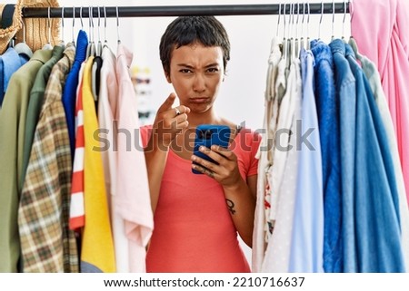Young hispanic woman with short hair searching clothes on clothing rack using smartphone pointing with finger to the camera and to you, confident gesture looking serious 